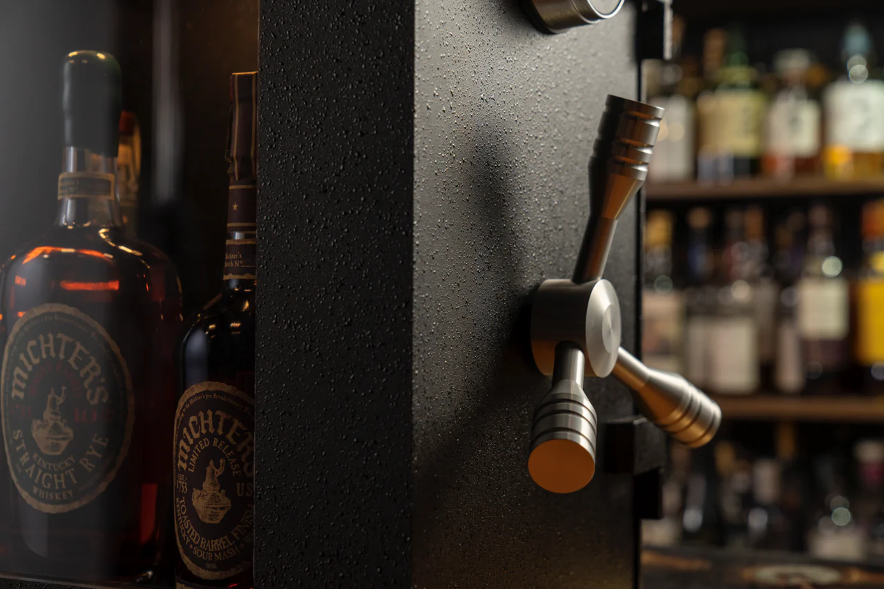 The Mini Vault is your ultimate sanctuary for prized spirits collections. Crafted with spirits enthusiasts in mind, this Mini Vault offers secure and stylish storage for 10-15 bottles of your finest spirits. The robust steel plate construction ensures durability and security, allowing you to rest easy knowing your valuable bottles are safe from unwanted access.
<br /><br />
  Designed to blend seamlessly into your space, the Mini Vault fits neatly on countertops or standalone bars, making it a convenient addition to any setting. The distinctive machined aluminum tri-spoke handle adds a touch of sophistication while providing easy access to your curated collection. The UL-certified La Gard Electronic Lock, paired with a bulletproof 19mm glass window, ensures both security and an impressive display of your spirits assortment, all while keeping it protected from unintended opening.
<br /><br />
  With its patented design and individually numbered for authenticity, the Mini Vault stands out as a piece of functional art. For those seeking a personal touch, custom airbrushed artwork on the inside of the door is available for an additional cost.
<br /><br />
  Whether you're a whiskey connoisseur or a business owner in the hospitality industry, this handcrafted masterpiece offers the perfect blend of security, style, and peace of mind. Elevate your personal bar experience - choose the Mini Vault, where craftsmanship meets security.