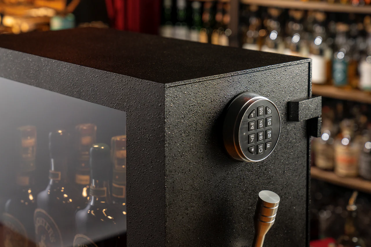 The Mini Vault is your ultimate sanctuary for prized spirits collections. Crafted with spirits enthusiasts in mind, this Mini Vault offers secure and stylish storage for 10-15 bottles of your finest spirits. The robust steel plate construction ensures durability and security, allowing you to rest easy knowing your valuable bottles are safe from unwanted access.
<br /><br />
  Designed to blend seamlessly into your space, the Mini Vault fits neatly on countertops or standalone bars, making it a convenient addition to any setting. The distinctive machined aluminum tri-spoke handle adds a touch of sophistication while providing easy access to your curated collection. The UL-certified La Gard Electronic Lock, paired with a bulletproof 19mm glass window, ensures both security and an impressive display of your spirits assortment, all while keeping it protected from unintended opening.
<br /><br />
  With its patented design and individually numbered for authenticity, the Mini Vault stands out as a piece of functional art. For those seeking a personal touch, custom airbrushed artwork on the inside of the door is available for an additional cost.
<br /><br />
  Whether you're a whiskey connoisseur or a business owner in the hospitality industry, this handcrafted masterpiece offers the perfect blend of security, style, and peace of mind. Elevate your personal bar experience - choose the Mini Vault, where craftsmanship meets security.